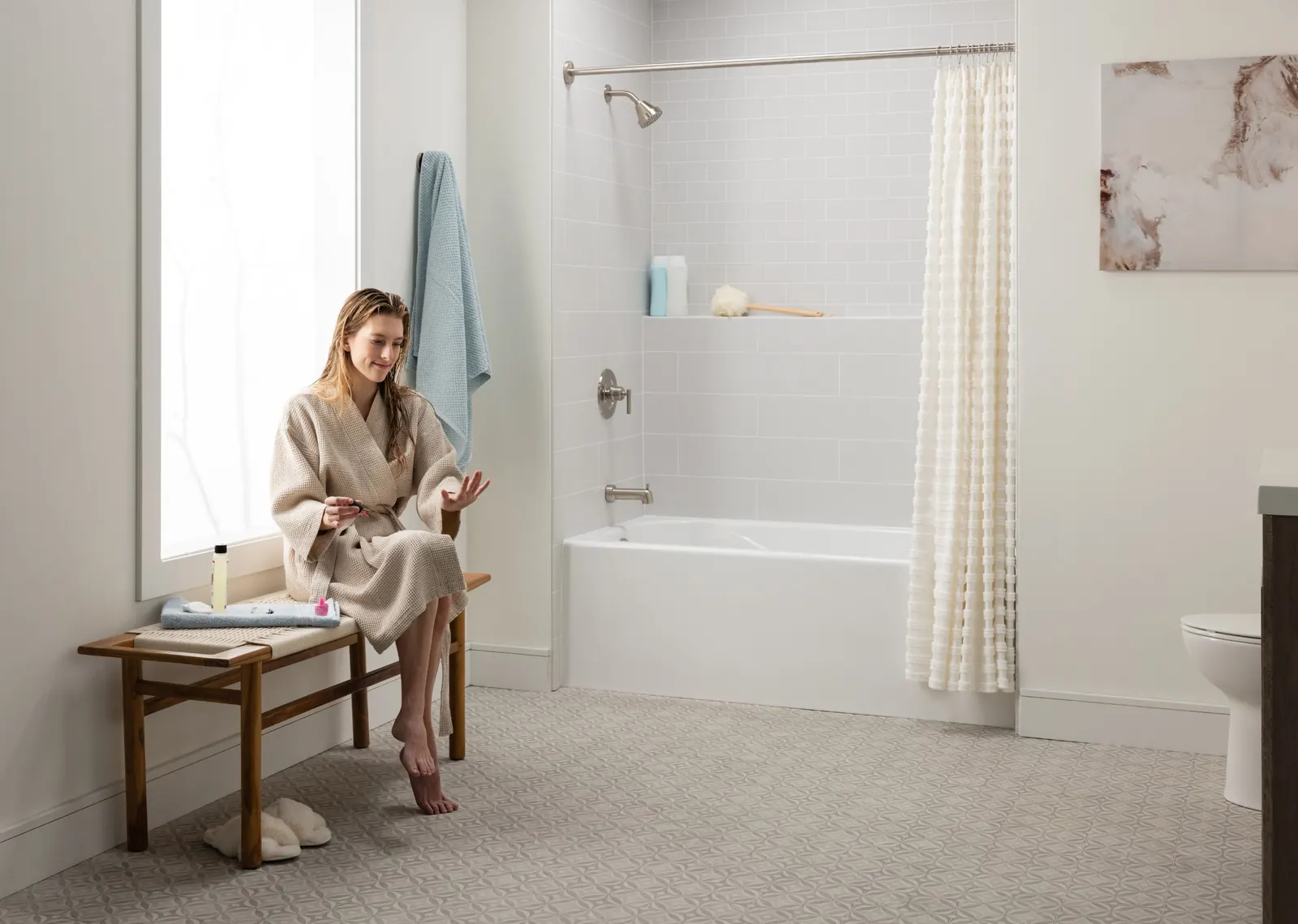 Woman in a robe sitting on a bench next to a white bathtub with light gray subway-pattern wall panels and a built-in shelf