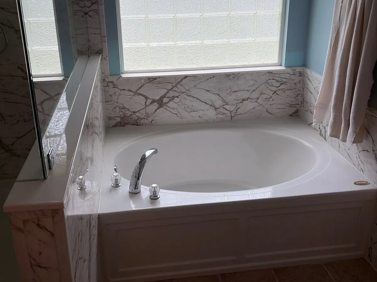 White soaking tub with a paneled front, set in white marble-look grout-free wall panels under a glass block window
