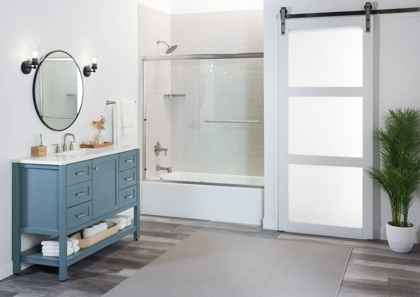 White tub and shower combo with light gray subway-pattern wall panels and a sliding glass door, next to a blue vanity