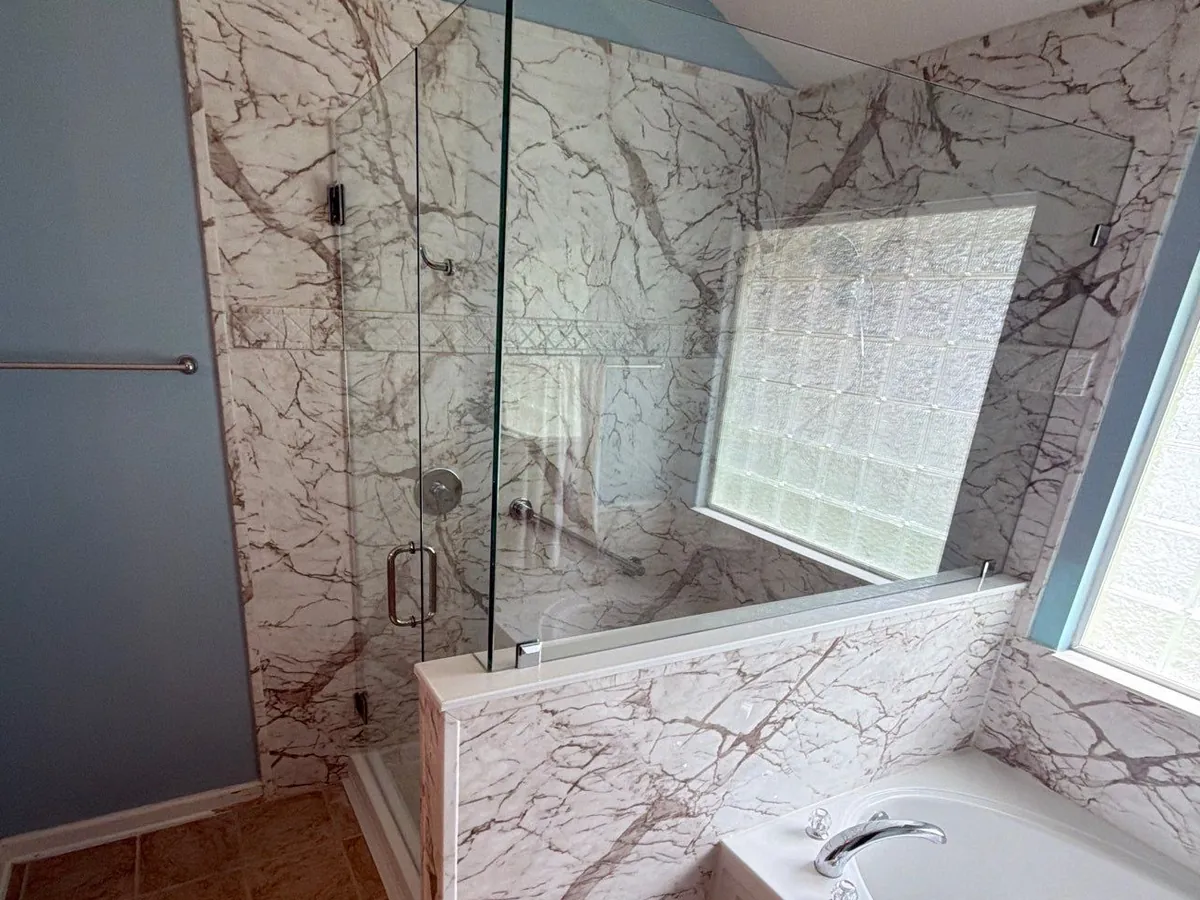 White marble-look grout-free wall panels in a glass-enclosed shower and around the soaking tub beside it, with a half wall and glass block window
