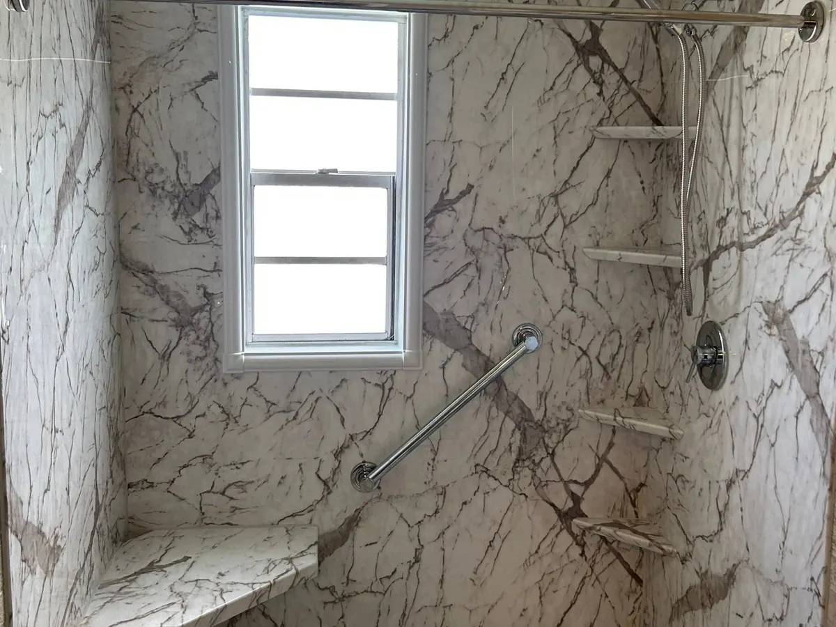 Walk-in shower with white marble-look grout-free wall panels, a corner seat, grab bar, corner shelves and a window