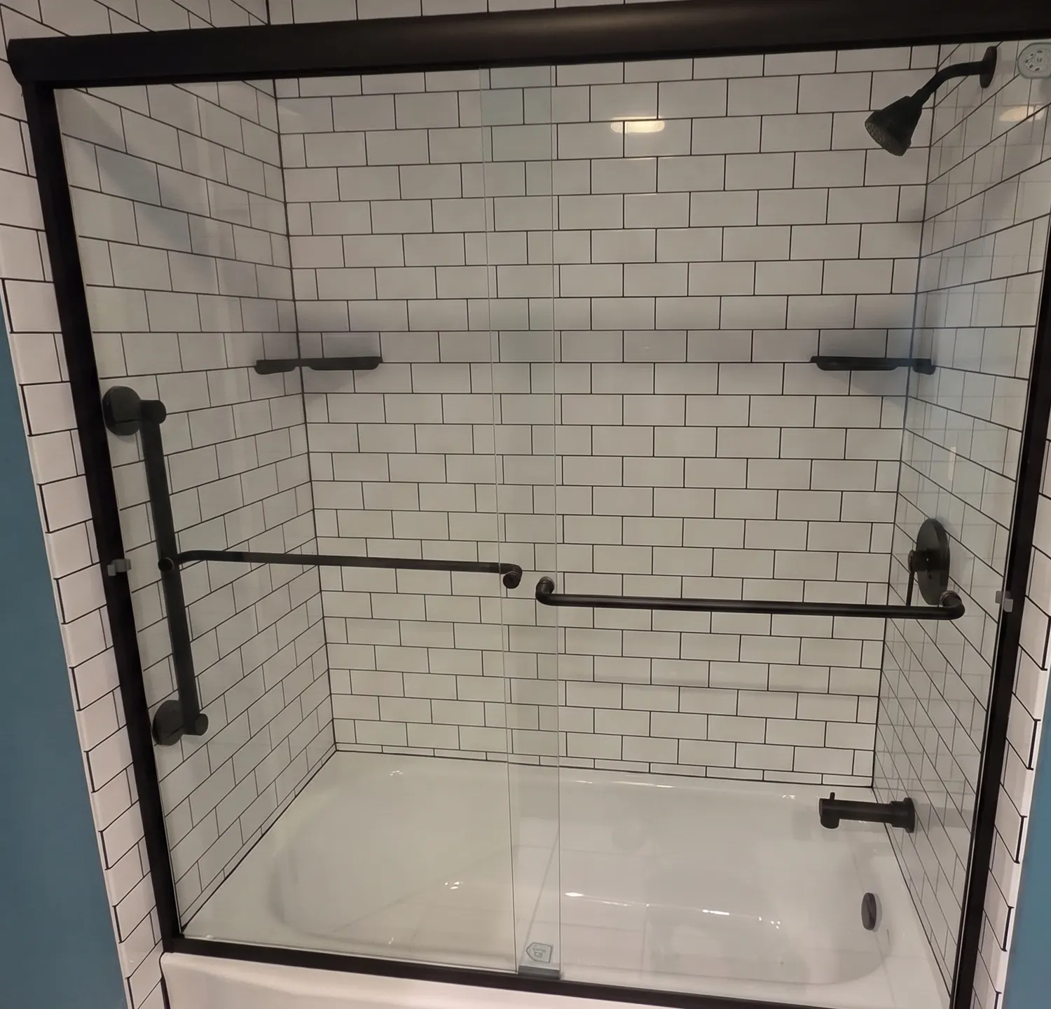 Tub and shower with white subway-pattern grout-free wall panels, matte black sliding glass door, grab bar, shelves and fixtures