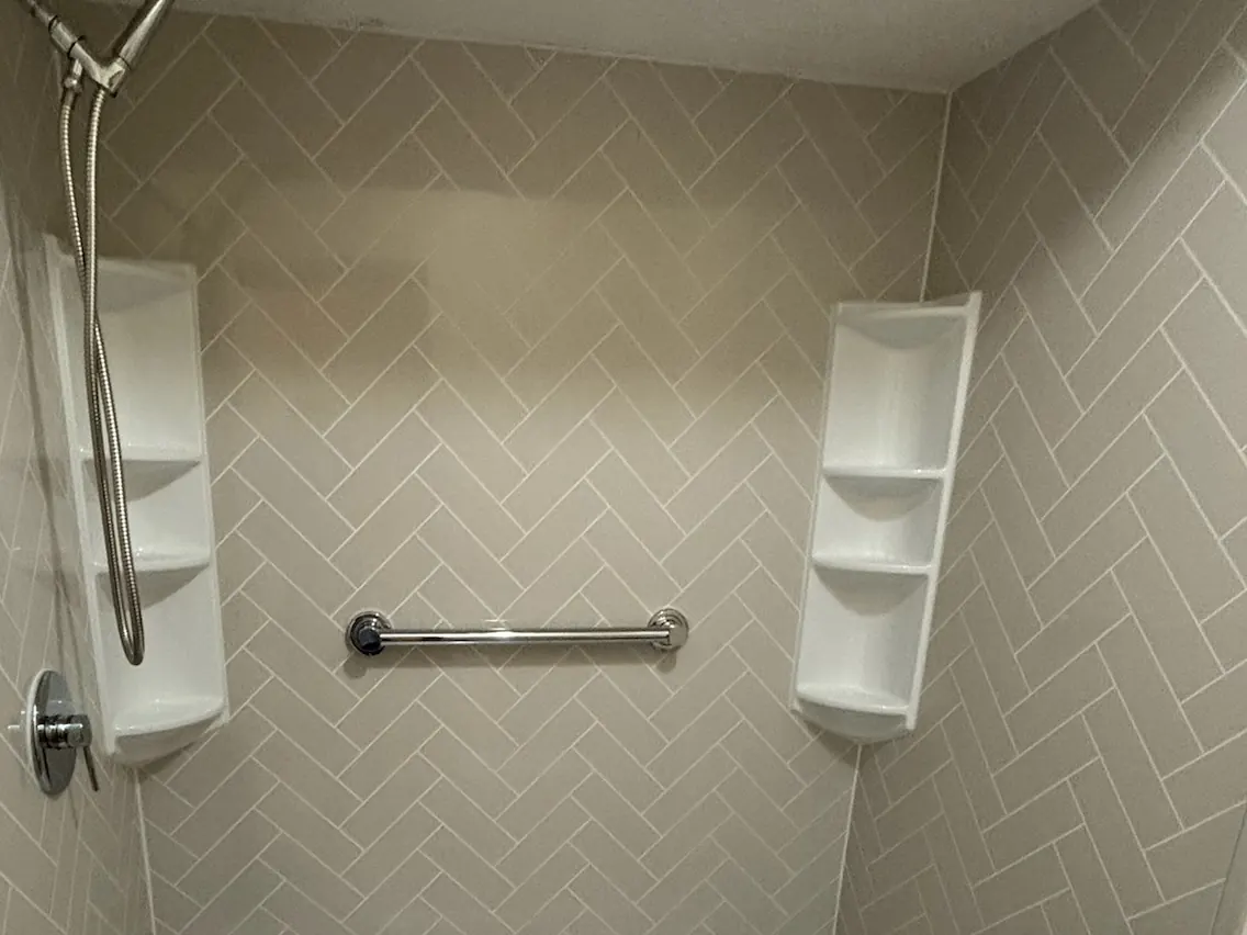 Shower with greige herringbone-pattern grout-free wall panels, white corner shelves, a grab bar and a handheld showerhead