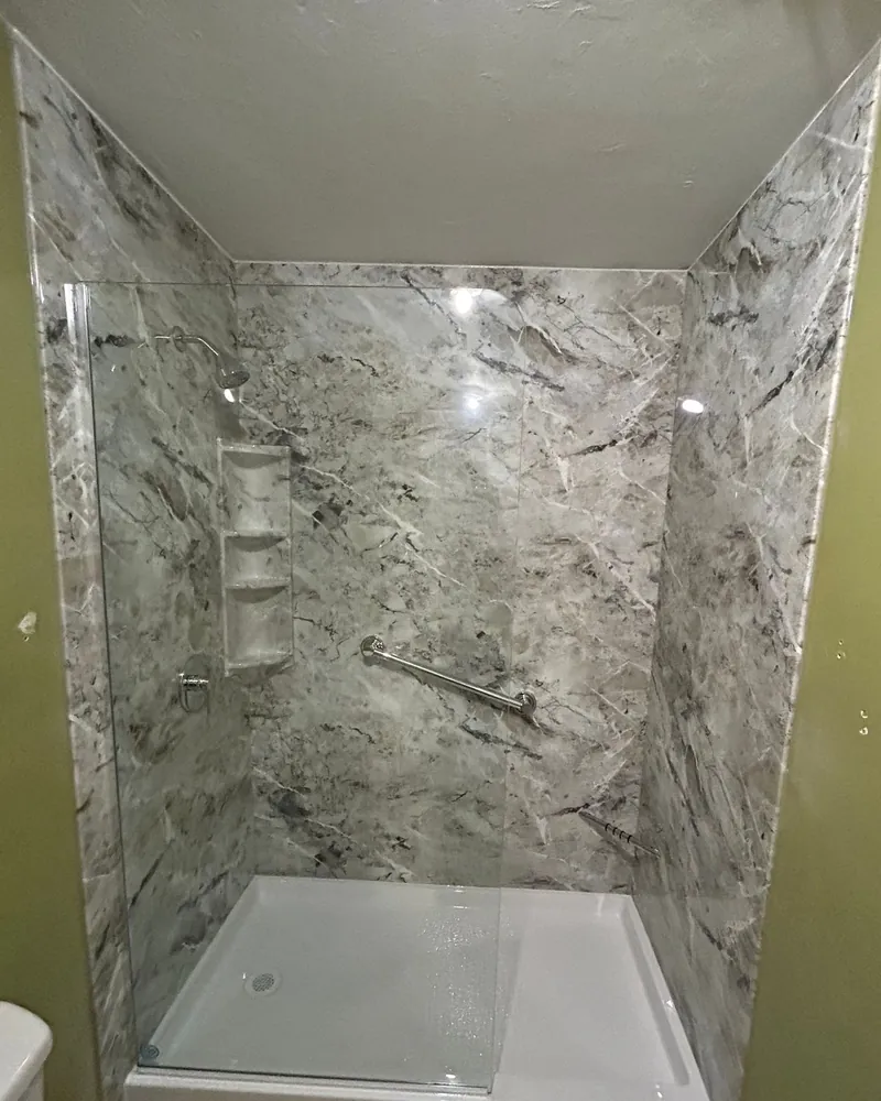 After: walk-in shower with gray marble-look wall panels, a white shower base, corner shelves, a grab bar and a clear glass panel