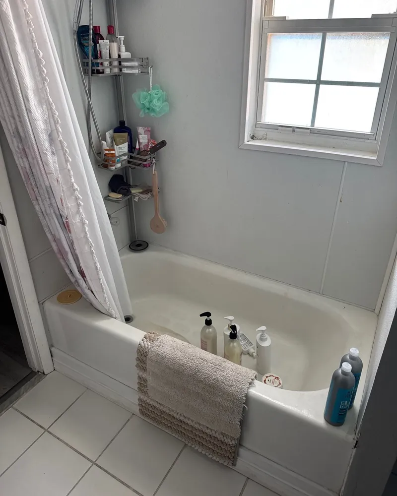 Before: worn white tub with a shower curtain, a pole caddy full of bottles and plain white wall panels under a window