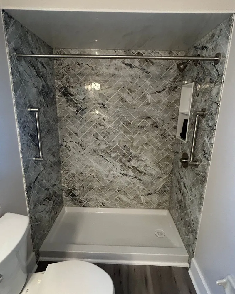 After: walk-in shower with gray marble-look herringbone wall panels, a built-in niche, two grab bars and a low white shower base