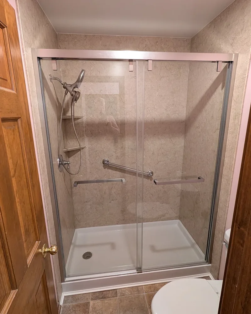 Walk-in shower with tan stone-look wall panels, a low white base, a sliding glass door, a grab bar, corner shelves and a handheld showerhead