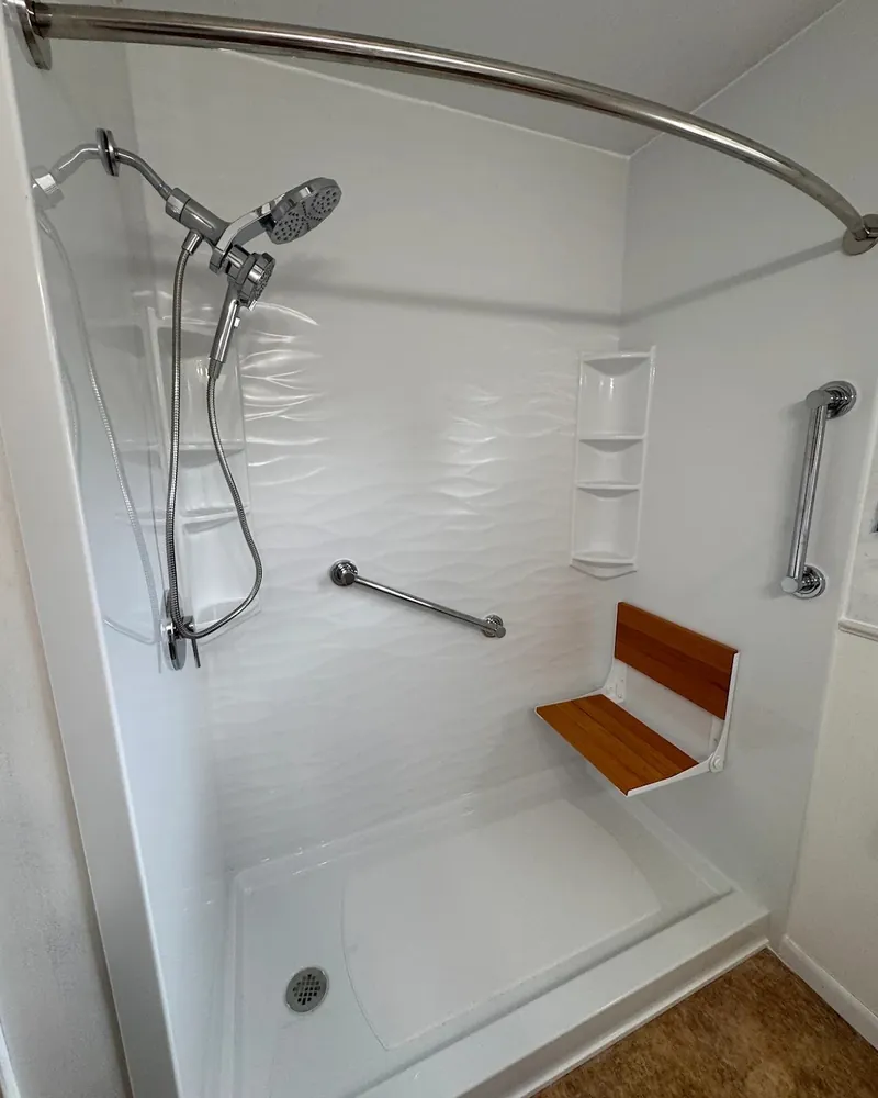 Walk-in shower with white wave-pattern wall panels, a low white base, a fold-down teak seat, two grab bars, corner shelves and a handheld showerhead