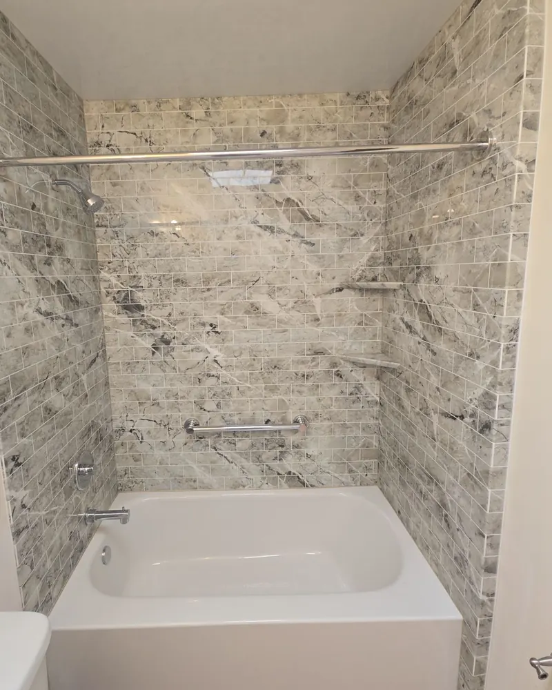 new white tub with gray marble-look subway-pattern wall panels, corner shelves and a grab bar