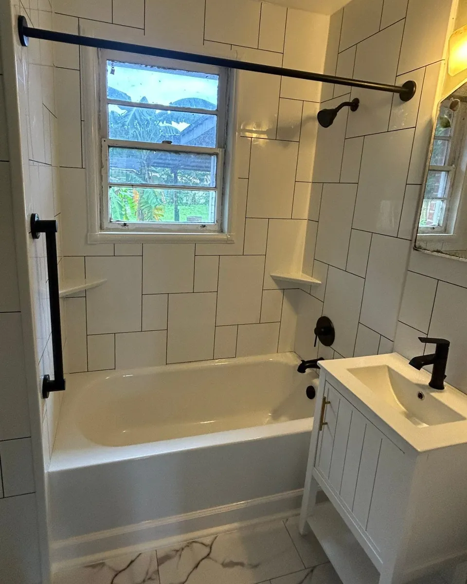 New white bathtub with white tile-look grout-free wall panels, two corner shelves, a matte black grab bar, showerhead and tub faucet under a window, with a new white vanity and marble-look floor