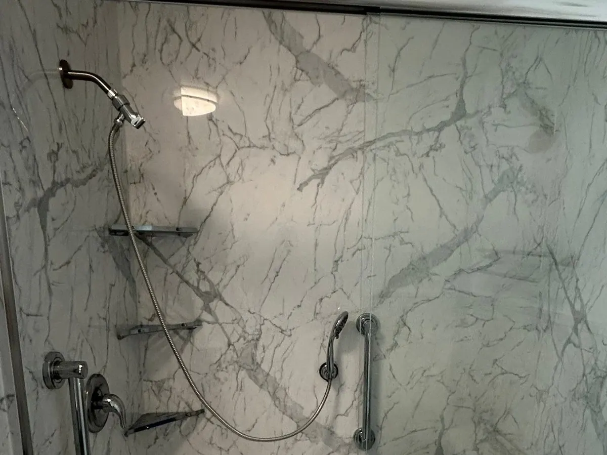 Shower with white marble-look grout-free wall panels, two grab bars, corner shelves, a handheld showerhead and a single-handle valve