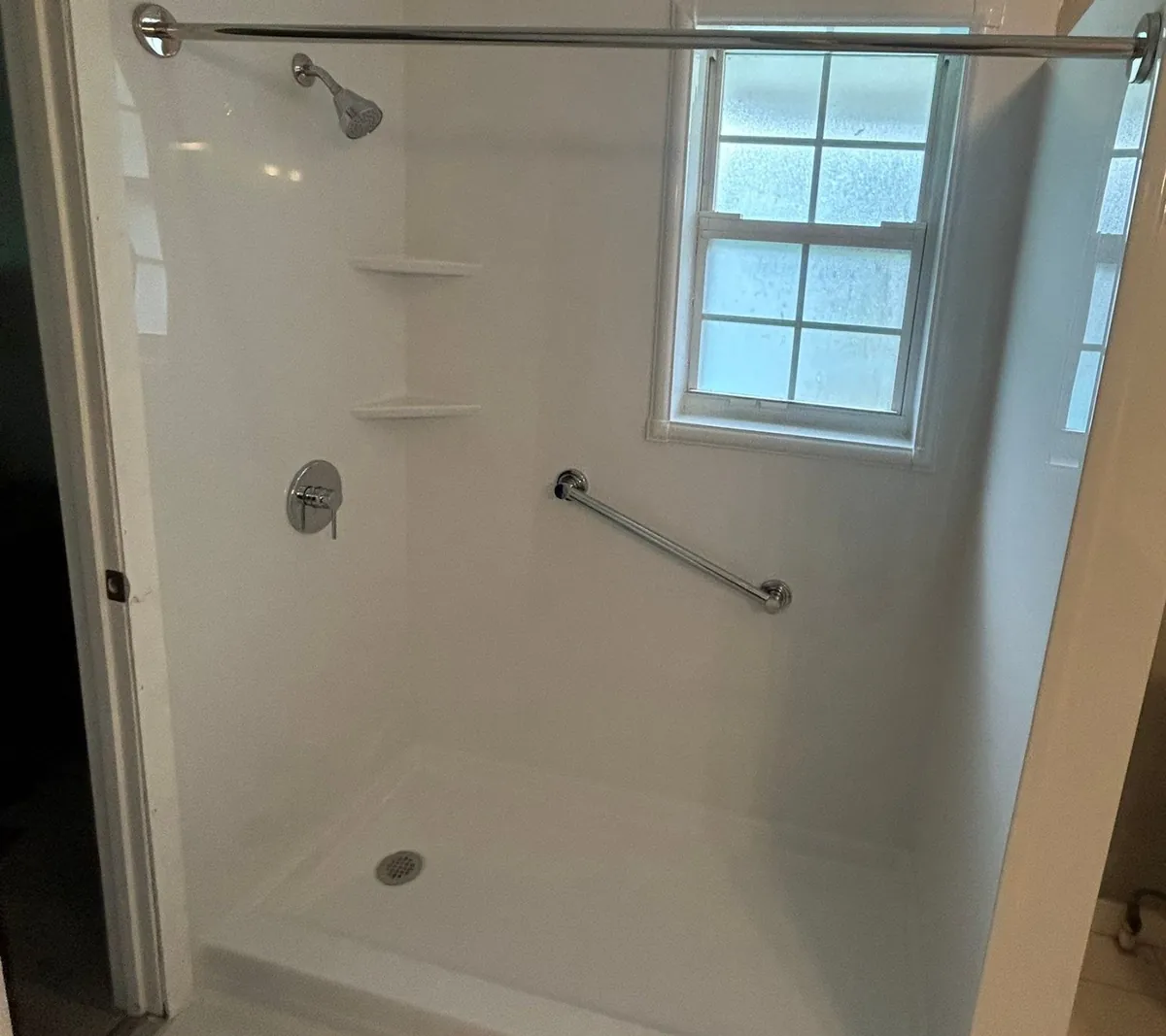 Finished walk-in shower with white grout-free walls, a low shower base, two corner shelves, a chrome valve, an angled grab bar, a window and a curtain rod